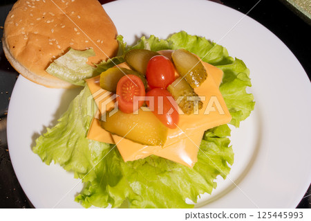 An open-faced burger with a sesame bun, lettuce, cheese, pickles, and cherry tomatoes on a white plate, highlighting vibrant burger preparation. 125445993