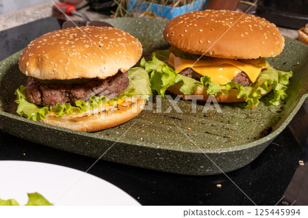 Two burgers with sesame buns, beef patties, lettuce, melted cheese, and sauce on a green grill pan, perfect for homemade meal scenes. Two burgers with sesame buns, beef patties, lettuce, melted cheese, and sauce on a green grill pan, perfect for homemade meal scenes. 125445994