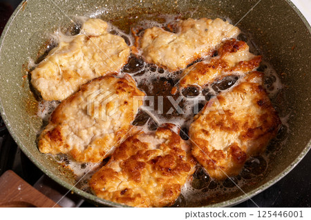 Four golden-brown breaded chicken pieces sizzling in bubbling oil inside a pan with a speckled surface. 125446001
