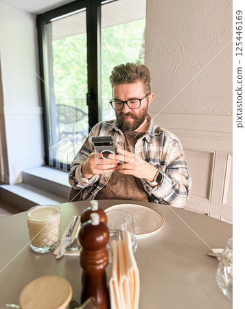 Handsome man browses his smartphone while waiting for coffee. Digital routine, casual lifestyle and urban cafe culture 125446169