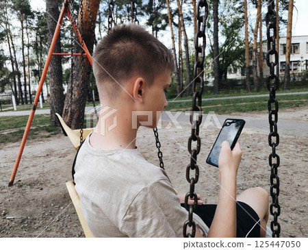 A young guy uses a smartphone outdoors while sitting on a swing in the park. The concept of social media and addiction. Teenagers' dependence on gadgets. A young guy uses a smartphone outdoors while sitting on a swing in the park. The concept of social media and addiction. Teenagers' dependence on gadgets. 125447050