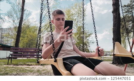 A young guy uses a smartphone outdoors while sitting on a swing in the park. The concept of social media and addiction. Teenagers' dependence on gadgets. 125447051