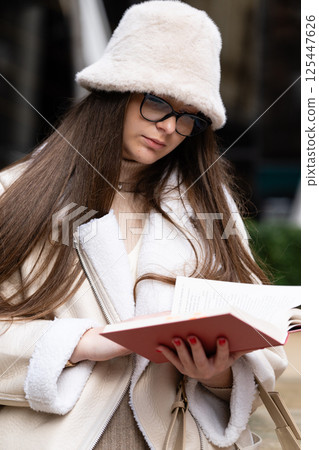 Reading quietly in a cozy outfit on a bustling city street as the sun sets, a young woman immerses herself in a book while dressed for warmth and style Reading quietly in a cozy outfit on a bustling city street as the sun sets, a young woman immerses herself in a book while dressed for warmth and style 125447626