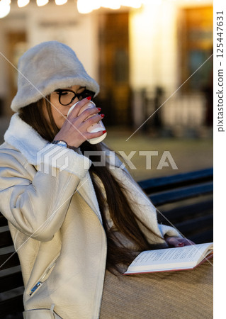 Cozy evening with a warm drink and a good book while sitting on a park bench under soft glowing lights in a charming urban setting Cozy evening with a warm drink and a good book while sitting on a park bench under soft glowing lights in a charming urban setting 125447631