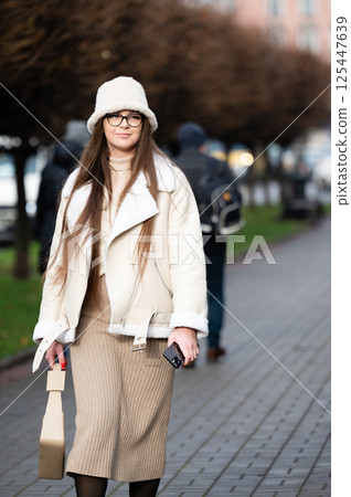 A fashionable woman, dressed in a snug and cozy outfit, strolls gracefully along the city sidewalk, enveloped by vibrant autumn trees on a pleasantly cool day A fashionable woman, dressed in a snug and cozy outfit, strolls gracefully along the city sidewalk, enveloped by vibrant autumn trees on a pleasantly cool day 125447639