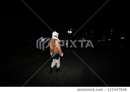 Exploring a quiet urban night wearing stylish layers and a white hood, a figure walks down a cobblestone path illuminated by soft streetlights in the city 125447646