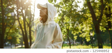 Young woman in a white hoodie and cap standing in a park on a sunny day. 125448952