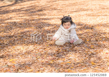 [One-year-old child sitting and playing in a winter park covered with fallen leaves] 125452069