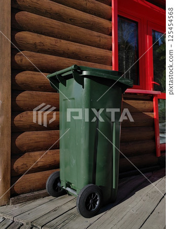 green trash bin for recycling positioned near wooden cabin with bright red window. green trash bin for recycling positioned near wooden cabin with bright red window. 125454568