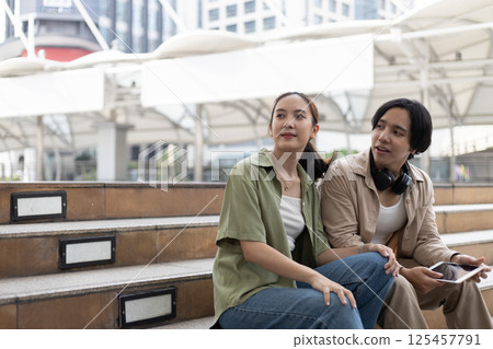 Two young adult engage in a relaxed conversation on stepped seating in a contemporary urban environment, surrounded by buildings and natural light during the day. 125457791