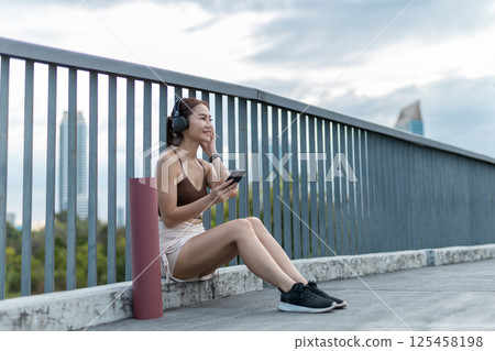 Young beautiful women sportswear with yoga mat smartphone and listening to music with headphones relax in park during exercise.fitness healthy concept. Young beautiful women sportswear with yoga mat smartphone and listening to music with headphones relax in park during exercise.fitness healthy concept. 125458198