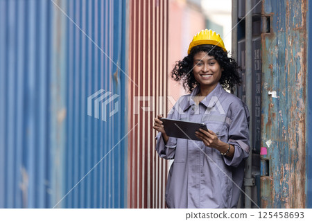 Female African American smiling engineering in uniform wear hard hat standing containers yard holding tablet. Area logistics import export and shipping. Female African American smiling engineering in uniform wear hard hat standing containers yard holding tablet. Area logistics import export and shipping. 125458693