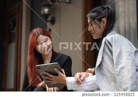 Two women sitting using tablet presenting work and discussing ideas outdoor environment during afternoon. Their engaged expressions suggest meaningful dialogue. female colleague brainstorming working. Two women sitting using tablet presenting work and discussing ideas outdoor environment during afternoon. Their engaged expressions suggest meaningful dialogue. female colleague brainstorming working. 125458825