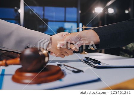 Two businesspeople are shaking hands in a law office with a scale and a gavel on the table 125461281