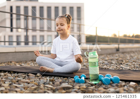 Outdoor scene of young girl sitting in lotus position atop rocky surface meditating. She is wearing casual clothes with water bottle and weights nearby. Peaceful moment of mindfulness and relaxation. Outdoor scene of young girl sitting in lotus position atop rocky surface meditating. She is wearing casual clothes with water bottle and weights nearby. Peaceful moment of mindfulness and relaxation. 125461335