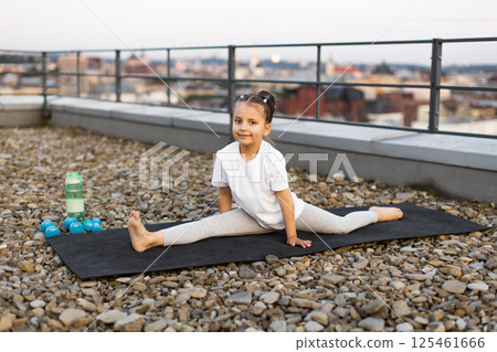 Young girl aged approximately seven years sitting in split pose on rooftop mat. Next to her are exercise equipment, representing activity, yoga, fitness training and outdoor healthy lifestyle 125461666