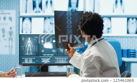 Physician reviewing MRI scan and x ray results with a patient in medical office, explaining the diagnostics. Doctor pointing at analysis to discuss about the illness, medical technology. Camera A. 125461869