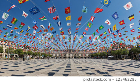 A large open square is decorated with numerous colorful flags strung on lines across the sky. Trees and buildings surround the square, creating a festive atmosphere 125462283