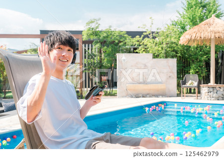 Young Asian man sitting on a recliner by the pool and playing games Young Asian man sitting on a recliner by the pool and playing games 125462979