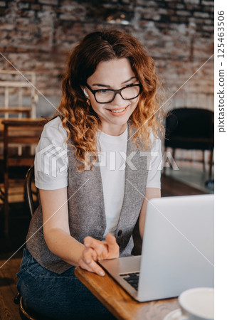 A young woman with curly red hair smiles while engaging in a video call from a cafe. Remote work and digital collaboration, hybrid workspace, online meeting, flexible work... A young woman with curly red hair smiles while engaging in a video call from a cafe. Remote work and digital collaboration, hybrid workspace, online meeting, flexible work... 125463506