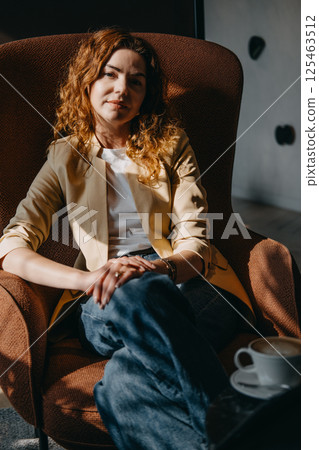 A confident young woman with curly red hair sits in a brown armchair, her hands folded in her lap. Strategic mindset, innovation leadership, poised professional, impactful women... 125463512