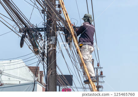Utility worker repairs power lines using a bamboo ladder in a vibrant urban setting in Thailand Utility worker repairs power lines using a bamboo ladder in a vibrant urban setting in Thailand 125465760