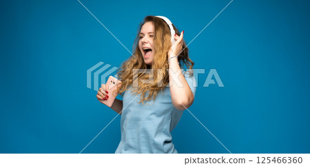 Happy young woman enjoying music in headphones with smartphone in hand on blue backdrop 125466360