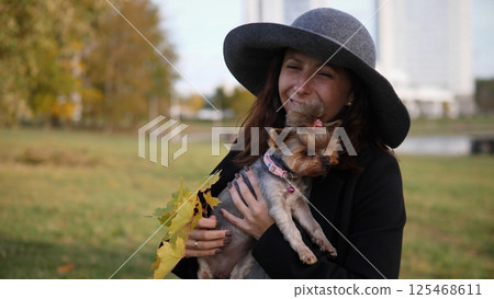 A joyful woman wearing a stylish hat holds her small dog close amidst the beautiful autumn foliage and scenery 125468611