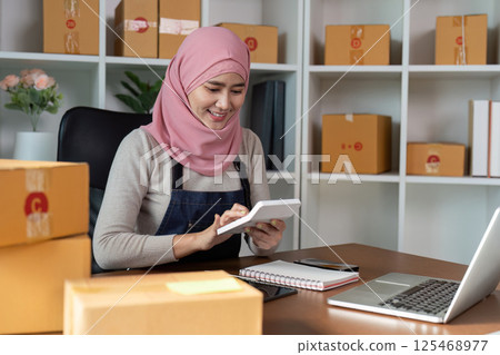 Happy Muslim woman checking her online business inventory and preparing packages in a modern office environment. 125468977