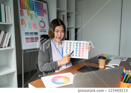 Graphic designer showcasing color palette options at her desk while engaging with a laptop in a modern workspace 125469215