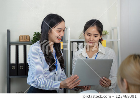 Enthusiastic Asian woman sharing insights from a laptop with a colleague during a collaborative meeting in a bright office 125469391