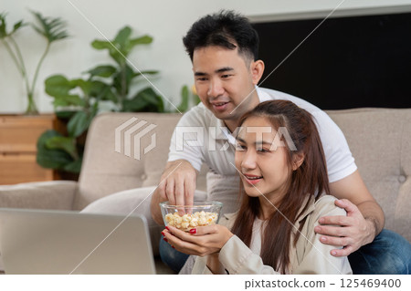 Couple Enjoying Movies and Snack Sharing. A couple sharing popcorn while watching a movie on their laptop at home. 125469400