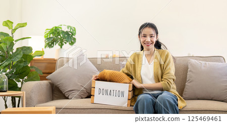 Smiling woman sitting on the couch with a donation box, proudly presenting folded clothes in a cozy living room 125469461
