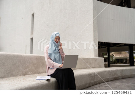 Muslim woman in hijab on a business call while working on her laptop Muslim woman in hijab on a business call while working on her laptop 125469481