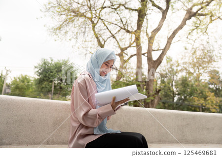 Muslim woman reviewing business documents in a serene outdoor setting. Muslim woman reviewing business documents in a serene outdoor setting. 125469681