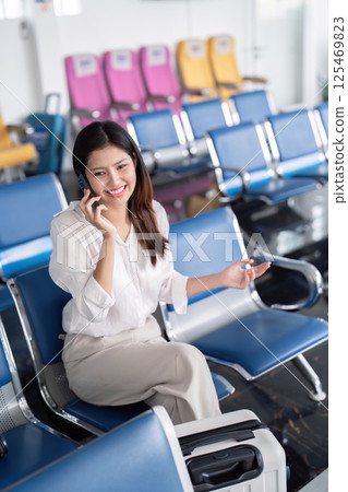 Smiling young woman talking on the phone while waiting in airport lounge with suitcase Smiling young woman talking on the phone while waiting in airport lounge with suitcase 125469823