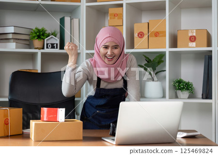 Muslim woman cheering for her online business success in a cozy workspace. 125469824