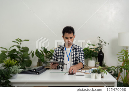 Remote work setup with young Asian man preparing materials in home office Remote work setup with young Asian man preparing materials in home office 125470068