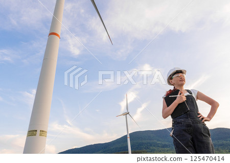 A confident female engineer proudly stands at a wind farm, overseeing the spinning turbines against a scenic backdrop, highlighting the promising future of sustainable energy solutions 125470414