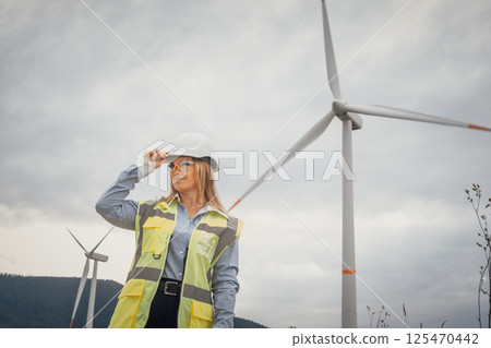 A highly confident female engineer, dressed in safety attire, stands proudly at a picturesque wind farm, showcasing her commitment to renewable energy and sustainable engineering practices 125470442