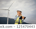 A confident female engineer in a safety vest and helmet analyzes data on her tablet at a wind farm, surrounded by wind turbines and natural scenery that highlights sustainable energy 125470443