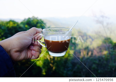 Hand holding a glass cup of hot coffee on the valley mountain in natural sunlight of morning bright day Hand holding a glass cup of hot coffee on the valley mountain in natural sunlight of morning bright day 125470956