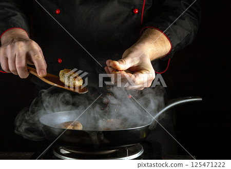 A chef skillfully cooks meatballs in a pan, adding spices to enhance flavor, surrounded by swirling steam in a dark kitchen setting 125471222