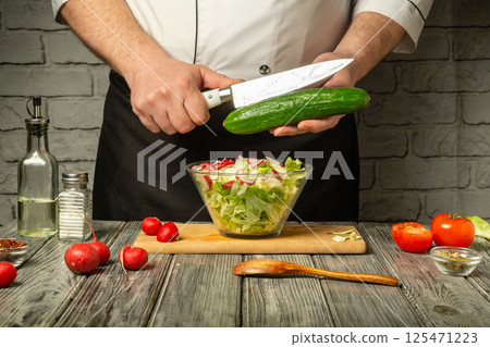 In a cozy kitchen, a chef skillfully slices a cucumber above a bowl of fresh salad. Colorful tomatoes and radishes enhance the vibrant display while seasonings await 125471223