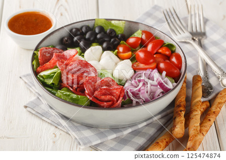 Salad with salami sausages, mozzarella cheese, tomatoes, olives, onions, romaine lettuce and spicy vinaigrette dressing served with breadsticks close-up in a bowl. Horizontal 125474874