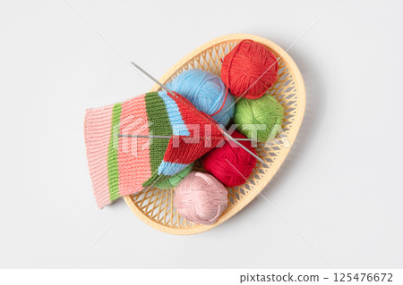 Red, green, blue and pink knitting balls in basket with knitting needles and knitted scarf on gray background. Set for the hobby of knitting warm clothes or toys. Concept needlework Red, green, blue and pink knitting balls in basket with knitting needles and knitted scarf on gray background. Set for the hobby of knitting warm clothes or toys. Concept needlework 125476672