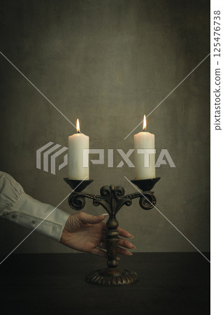 Human hand with sharp nails in white vintage shirts sleeve touches old brass candlestick with burning candles in the dark, vertical image. 125476738