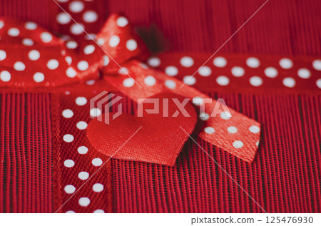 Close up of a red tiny heart and gift ribbon with white polka dots tied into a bow, placed on a textured red and pink striped background 125476930