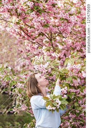 Smiling woman standing among blooming pink cherry blossom sakura trees wearing a light blue shirt and enjoying a bright spring day outdoors 125479089