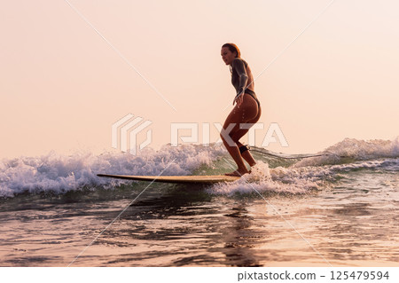 Surfer on surfboard rides on wave in morning with warm sunrise. Woman riding on longboard. Surfer on surfboard rides on wave in morning with warm sunrise. Woman riding on longboard. 125479594